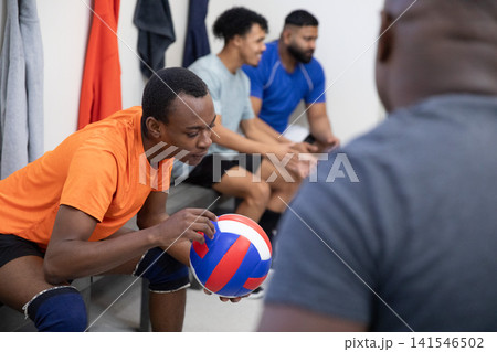 African American male teens sitting on bench in locker room, orange-shirt teen holding volleyball African American male teens sitting on bench in locker room, orange-shirt teen holding volleyball 141546502
