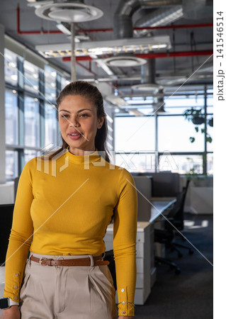 Woman speaking near desks in open office wearing mustard top, beige trousers, smartwatch Woman speaking near desks in open office wearing mustard top, beige trousers, smartwatch 141546514