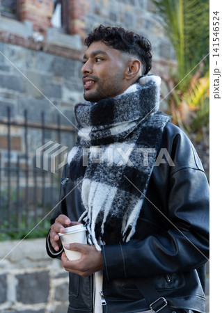 Indian man standing on sidewalk by stone wall, wearing leather jacket and checked scarf holding cup Indian man standing on sidewalk by stone wall, wearing leather jacket and checked scarf holding cup 141546524