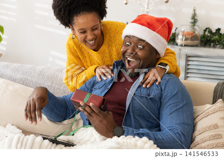 African American couple sitting on living room sofa, man holding wrapped gift, wearing Santa hat 141546533
