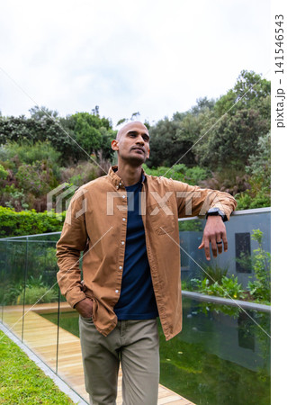 Asian man leaning on glass railing along boardwalk in garden beside pool, wearing smartwatch Asian man leaning on glass railing along boardwalk in garden beside pool, wearing smartwatch 141546543