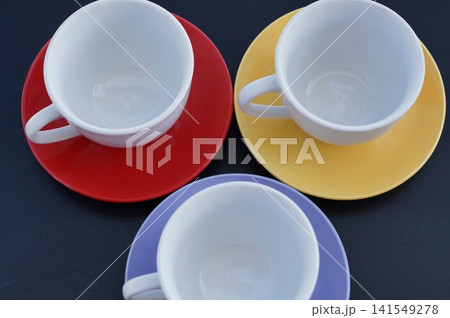 Three white ceramic cups are arranged on red, yellow, and purple saucers in a top view. The colorful table setting represents friendship, shared drinks, and welcoming home traditions Three white ceramic cups are arranged on red, yellow, and purple saucers in a top view. The colorful table setting represents friendship, shared drinks, and welcoming home traditions 141549278