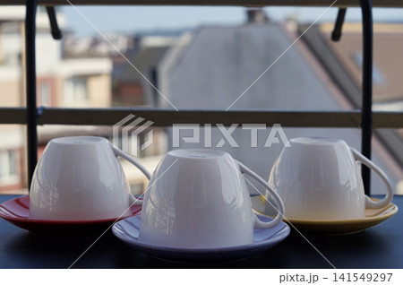 Three inverted white cups are displayed on colorful saucers against a softly blurred city background. The still life expresses domestic order, simplicity, and anticipation of a shared drink Three inverted white cups are displayed on colorful saucers against a softly blurred city background. The still life expresses domestic order, simplicity, and anticipation of a shared drink 141549297