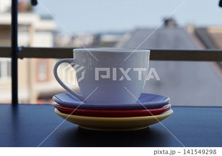 A white ceramic cup rests on a stack of purple, red, and yellow saucers near a balcony window. The colorful still life symbolizes simple morning rituals, comfort, and cheerful home hospitality 141549298