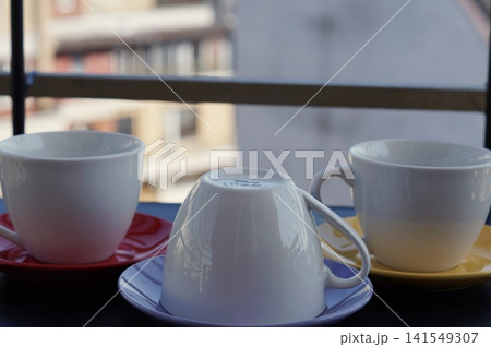 White cups and colorful saucers are arranged on a balcony table against a blurred city backdrop. The still life expresses hospitality, simplicity, and cozy home living. 141549307