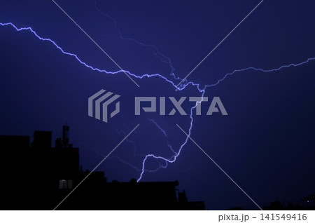 Bright branching lightning illuminates a dark blue night sky above silhouetted city buildings. Dramatic thunderstorm scene represents natural power, danger, and fleeting energy. 141549416