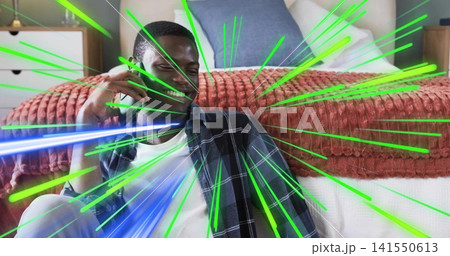 Smiling man holding smartphone, sitting on floor by bed, wearing plaid shirt under green-blue glow Smiling man holding smartphone, sitting on floor by bed, wearing plaid shirt under green-blue glow 141550613