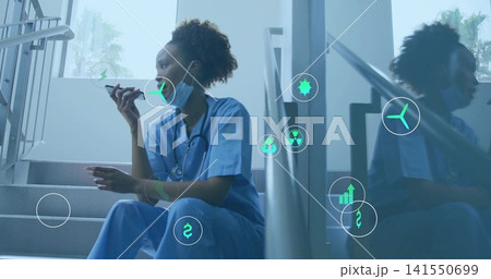 Sitting nurse in blue scrubs speaking into phone on stairwell, with surgical mask lowered, AR icons Sitting nurse in blue scrubs speaking into phone on stairwell, with surgical mask lowered, AR icons 141550699