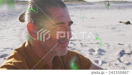 Gazing mature man sitting on sandy beach, brown shirt, tied-back bun with hair tie, green kite 141550867