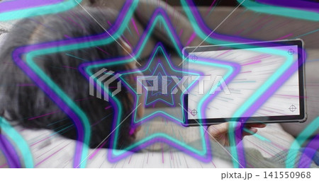 Reclining Chinese woman wearing sweater holding tablet in bedroom, with neon star graphic overlay 141550968