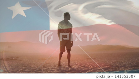 Standing silhouetted man wearing patterned shirt and shorts on sandy beach at sunset, flag overlay 141552307