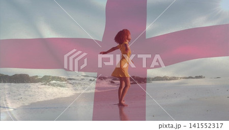 Standing woman in yellow sleeveless dress extending arms on sandy shoreline with pink cross overlay 141552317