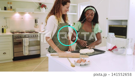 Two women wearing olive aprons kneading dough at white kitchen island, with rolling pin and eggs Two women wearing olive aprons kneading dough at white kitchen island, with rolling pin and eggs 141552658