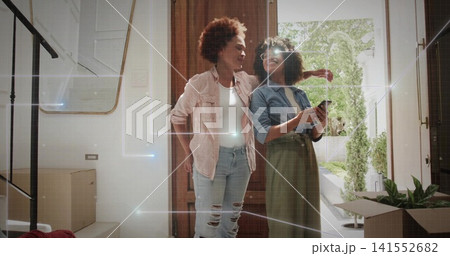 Standing two women embracing at home entryway, wearing denim jacket, holding smartphone near boxes Standing two women embracing at home entryway, wearing denim jacket, holding smartphone near boxes 141552682