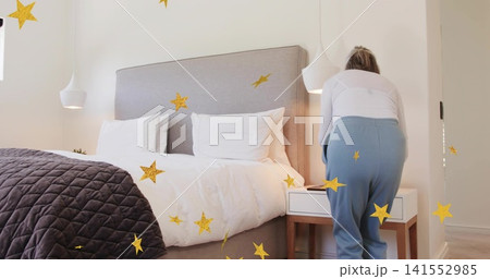 Bending senior woman in white top reaching phone on nightstand in bedroom, gold stars, copy space Bending senior woman in white top reaching phone on nightstand in bedroom, gold stars, copy space 141552985