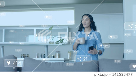 Standing woman in denim shirt holding smartphone and mug in kitchen with floating icons, copy space 141553477