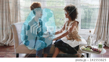 Holding hands, two women sleeveless top sitting on settee at home with fruit and hexagonal overlay Holding hands, two women sleeveless top sitting on settee at home with fruit and hexagonal overlay 141553825