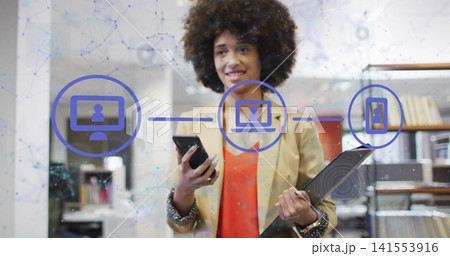 Standing woman wearing beige blazer in office, holding phone and tablet showing blue network icons Standing woman wearing beige blazer in office, holding phone and tablet showing blue network icons 141553916