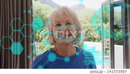 Standing senior woman in blue top and gold necklace at open glass door, teal holographic overlay Standing senior woman in blue top and gold necklace at open glass door, teal holographic overlay 141553997