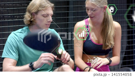 Adjusting athletic partners checking wrist devices in gym by grid wall with paddle and AR overlays Adjusting athletic partners checking wrist devices in gym by grid wall with paddle and AR overlays 141554058