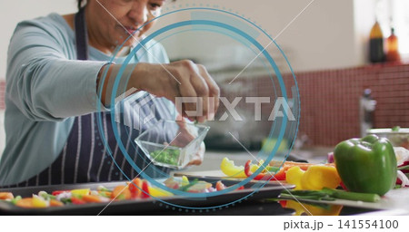 Sprinkling woman in striped apron, blue top at kitchen counter over baking trays with glass bowl 141554100