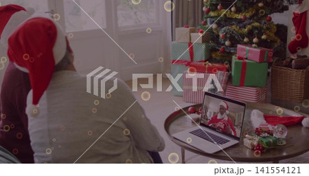Sitting couple wearing red-and-white Santa hats at home watching laptop caller with tree gifts 141554121