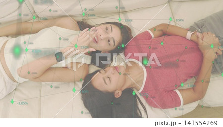 Lying two women talking on sofa cushions, wearing smartwatches, crop top, red tee, green overlay 141554269