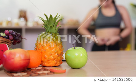 Fresh pineapple and green apple on table with woman measuring waist in background Fresh pineapple and green apple on table with woman measuring waist in background 141555500
