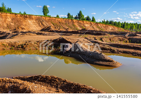 Scenic view of a post-mining sand quarry landscape featuring textured clay slopes, rugged earth formations, and calm muddy water reflections under a clear summer sky Scenic view of a post-mining sand quarry landscape featuring textured clay slopes, rugged earth formations, and calm muddy water reflections under a clear summer sky 141555580