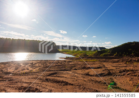 Sunny day at an abandoned sandy pit featuring a bright sun flare over a scenic lake, raw textured ground, and lush green forest silhouettes under a vast clear blue sky Sunny day at an abandoned sandy pit featuring a bright sun flare over a scenic lake, raw textured ground, and lush green forest silhouettes under a vast clear blue sky 141555585