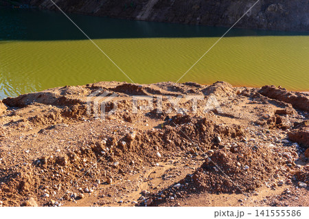 Rugged texture of sandy ground with scattered stones meeting the calm emerald waters of an abandoned quarry, illuminated by warm natural sunlight on a quiet summer day Rugged texture of sandy ground with scattered stones meeting the calm emerald waters of an abandoned quarry, illuminated by warm natural sunlight on a quiet summer day 141555586