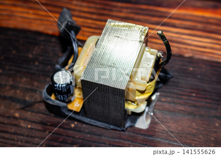 Disassembled electrical power supply unit revealing internal metal transformer and electronic components resting on a dark wooden surface, close-up view of broken electronic hardware parts 141555626