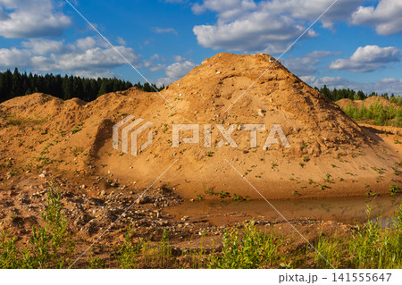 Large mound of yellow sand in an abandoned quarry with a small pool of water at the base, surrounded by wild vegetation under a bright blue sky with fluffy white clouds 141555647