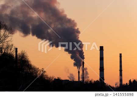 Factory smokestacks release thick dark plumes against a warm orange winter sunset, starkly silhouetted above bare trees 141555898