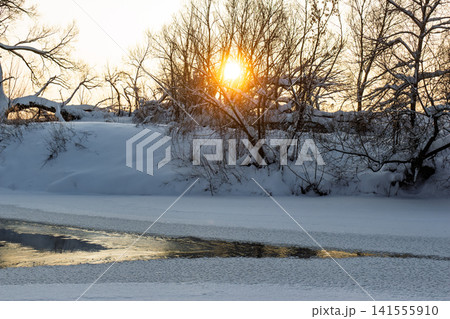 Brilliant winter sunset glowing through bare branches above a partially frozen river, creating a serene, frosty landscape scene 141555910