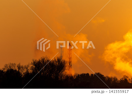 A fiery winter sunset illuminates a power plant chimney, as steam billows spectacularly against the stunning orange sky. Industrial beauty in a dramatic golden hour A fiery winter sunset illuminates a power plant chimney, as steam billows spectacularly against the stunning orange sky. Industrial beauty in a dramatic golden hour 141555917