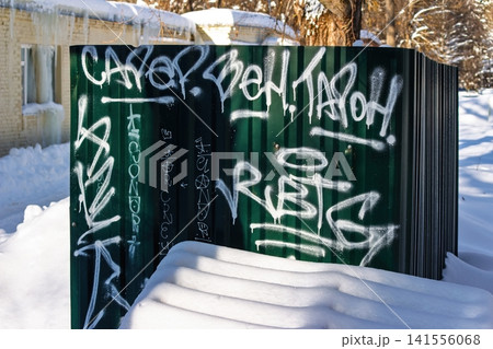 Bold white graffiti tags a dark green corrugated fence, blanketed by crisp winter snow. Urban street art expression in a frosty setting: Russia - January 30, 2026 Bold white graffiti tags a dark green corrugated fence, blanketed by crisp winter snow. Urban street art expression in a frosty setting: Russia - January 30, 2026 141556068