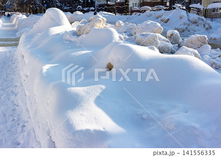 Freshly plowed snow, piled high along a clear winter sidewalk. Massive white snowdrifts create a stark contrast against the sunny urban background Freshly plowed snow, piled high along a clear winter sidewalk. Massive white snowdrifts create a stark contrast against the sunny urban background 141556335