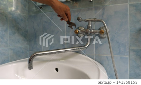 A man's hand uses an adjustable wrench to repair a faucet in a bathroom with blue tiles. The repairman tightens the fastening part on the metal faucet. The faucet body is partially disassembled. A man's hand uses an adjustable wrench to repair a faucet in a bathroom with blue tiles. The repairman tightens the fastening part on the metal faucet. The faucet body is partially disassembled. 141559155