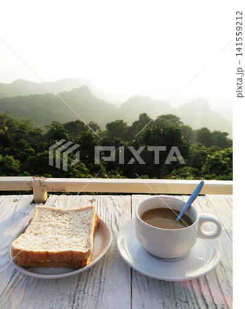 A cup of hot coffee with toast bread in white dish on white wooden counter bar is breakfast in resort on the mountain in Thailand A cup of hot coffee with toast bread in white dish on white wooden counter bar is breakfast in resort on the mountain in Thailand 141559212