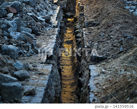 Sunlit narrow trench between stone rubble and excavated soil at construction site 141559516
