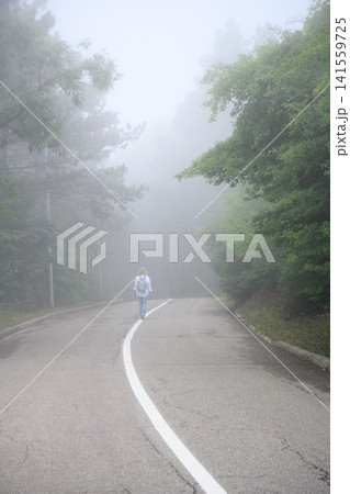 Vertical view of a woman walking on a foggy road. Lone traveler with backpack on a misty forest path. Journey and mystery concept. Copy space for text Vertical view of a woman walking on a foggy road. Lone traveler with backpack on a misty forest path. Journey and mystery concept. Copy space for text 141559725