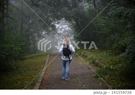 Rear view of a blonde woman walking on a path in a foggy forest. Female hiker with a black backpack exploring misty woods. Outdoor adventure, hiking and travel concept 141559726