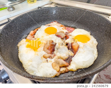 Fried eggs and bacon sizzling in a frying pan on a stove 141560679