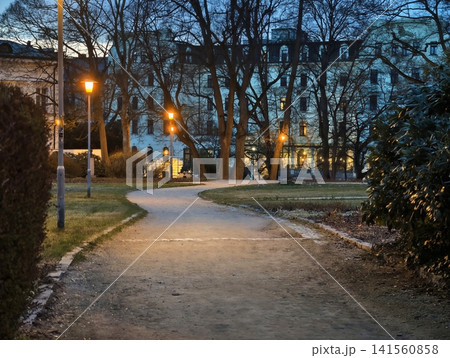 Peaceful park pathway with pedestrians resting beneath luminous amber lamps during calm blue evening Peaceful park pathway with pedestrians resting beneath luminous amber lamps during calm blue evening 141560858
