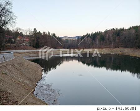 Serene lakeside with forest backdrop. Calm dusk by lake with reflective water and nearby houses Serene lakeside with forest backdrop. Calm dusk by lake with reflective water and nearby houses 141560881