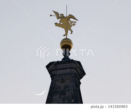 Golden angel silhouette. Decorative gilded angel atop stone column under crescent moonlit cloudy evening sky Golden angel silhouette. Decorative gilded angel atop stone column under crescent moonlit cloudy evening sky 141560963