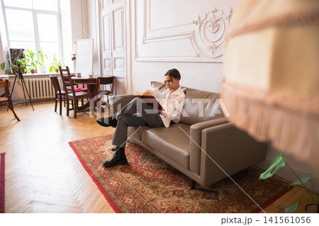 Man sitting on sofa reading book in cozy apartment. Stylish guy relaxing with literature in living room. Leisure time, comfortable lifestyle and intellectual hobby in home interior. 141561056