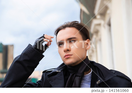Man in black trench coat wearing fingerless gloves looking away. Portrait of stylish young man outdoors. Fashion concept, urban street lifestyle, trendy male clothing and accessories. Man in black trench coat wearing fingerless gloves looking away. Portrait of stylish young man outdoors. Fashion concept, urban street lifestyle, trendy male clothing and accessories. 141561060