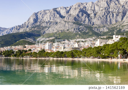 Makarska town and its vibrant beach line the Adriatic Sea, reflecting in the clear waters with the dramatic Biokovo Mountains creating a stunning backdrop Makarska town and its vibrant beach line the Adriatic Sea, reflecting in the clear waters with the dramatic Biokovo Mountains creating a stunning backdrop 141562189